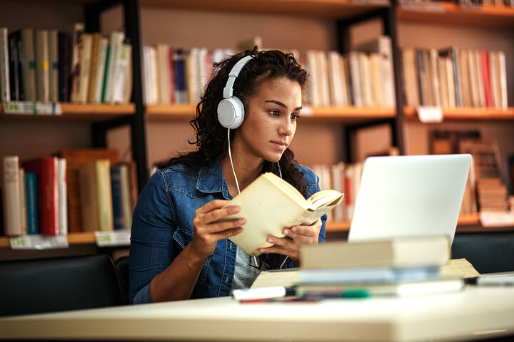 A student wearing headphones holds a book and looks at a laptop in a library.